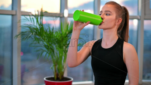 Video - Young woman drinking water after workout with city on background. Indoor training in apartment, quarantine