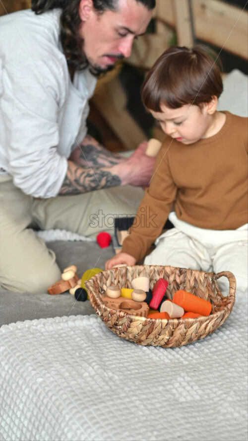 Video - Father playing with his son with colourful, ecological wooden toys from a basket on the bed