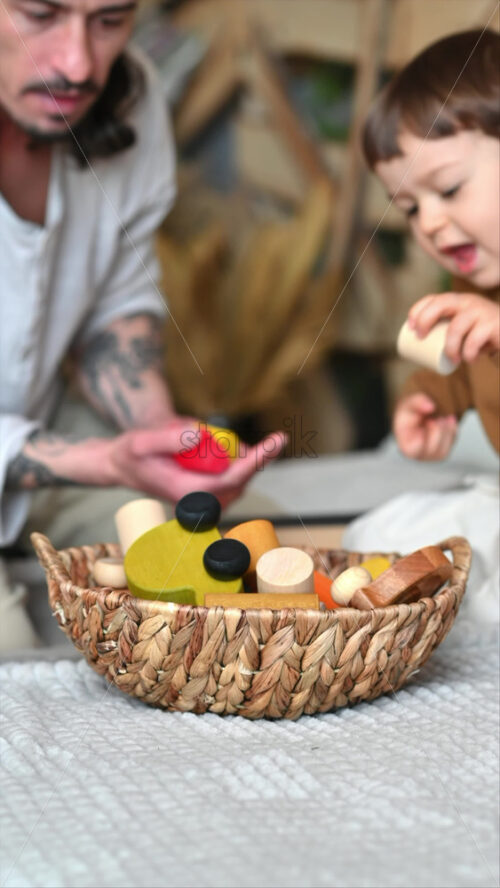 Video - Father playing with his son with colourful, ecological wooden toys from a basket on the bed. Vertical