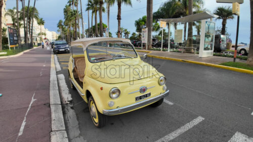 Video - Cannes, France - September 20, 2025: Vintage yellow Fiat 500 car parked on a palm lined street in the French Riviera