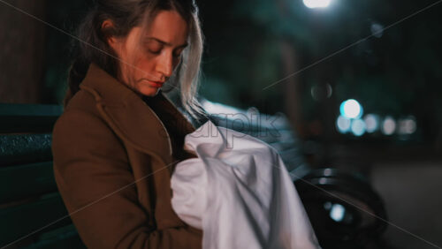 Video - A thoughtful woman resting on a park bench under soft night lights, wrapped in a coat and holding a baby