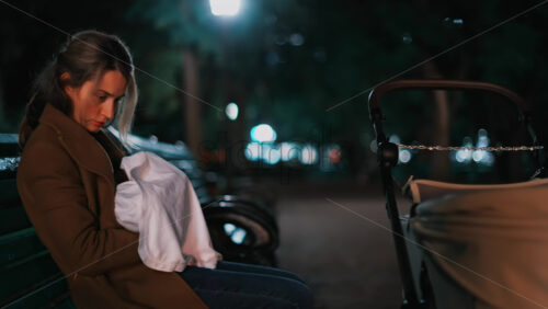 Video - A thoughtful woman resting on a park bench under soft night lights, wrapped in a coat and holding a baby