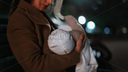 Video - A thoughtful woman resting on a park bench under soft night lights, wrapped in a coat and holding a baby