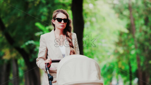 Video - Elegant woman in sunglasses walking confidently through a tree lined park with a beige stroller