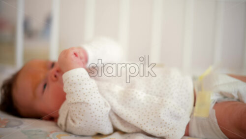 Video - A newborn baby in soft pajamas moves gently in a crib, stretching tiny hands in natural morning light