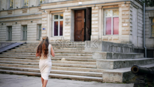 Video - Elegant woman with flowing hair and white dress walks up the stairs of a classic architectural building