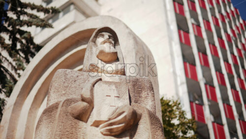 Video - Low angle view of a carved stone statue Petru Movila holding a book with a cross, framed by trees and modern architecture, Chisinau, Moldova