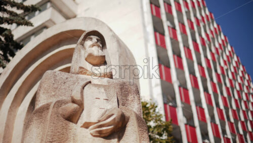 Video - Low angle view of a carved stone statue Petru Movila holding a book with a cross, framed by trees and modern architecture, Chisinau, Moldova