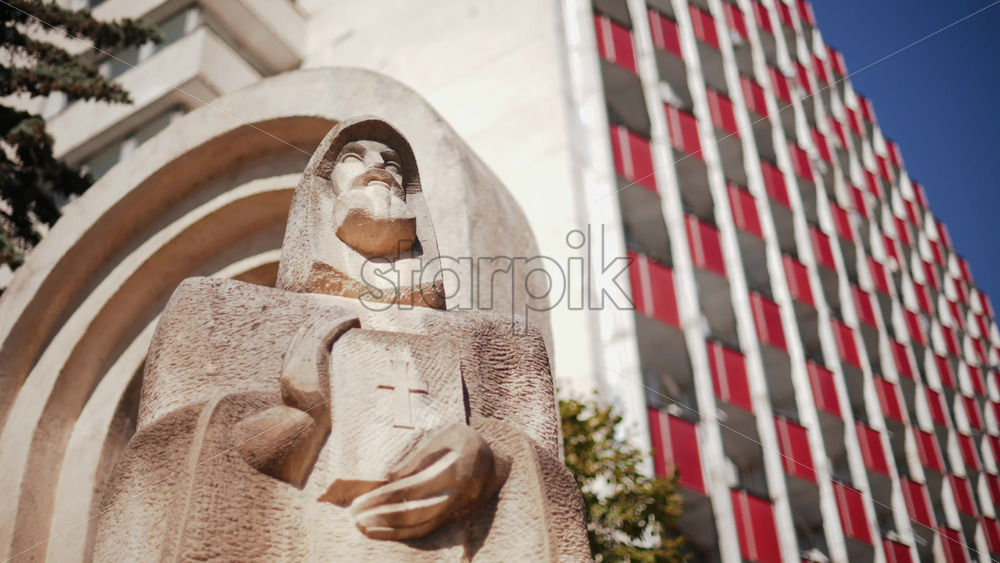 Video - Low angle view of a carved stone statue Petru Movila holding a book with a cross, framed by trees and modern architecture, Chisinau, Moldova