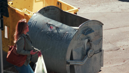 Video - Chisinau, Moldova - September 30, 2025: A woman holding her phone and bag throws a plastic trash bag into a large bin