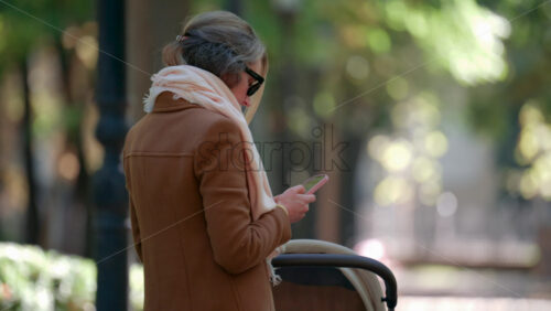 Video - A mother checking her phone while standing next to a baby carriage in the park