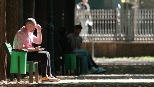Video - Chisinau, Moldova - September 30, 2025: Relaxed middle aged man resting on a bench, enjoying a warm day in a green urban park