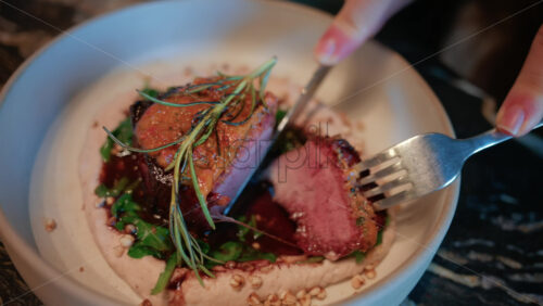 Video - Close up of a woman's hand cutting into a gourmet meat dish served with sauce, greens, and rosemary garnish on a white plate