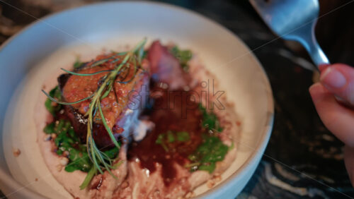 Video - Close up of a woman's hand cutting into a gourmet meat dish served with sauce, greens, and rosemary garnish on a white plate
