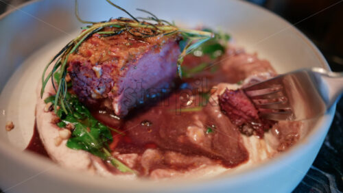 Video - Close up of a woman's hand cutting into a gourmet meat dish served with sauce, greens, and rosemary garnish on a white plate