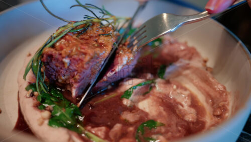 Video - Close up of a woman's hand cutting into a gourmet meat dish served with sauce, greens, and rosemary garnish on a white plate