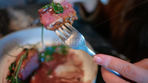 Video - Close up of a woman's hand cutting into a gourmet meat dish served with sauce, greens, and rosemary garnish on a white plate