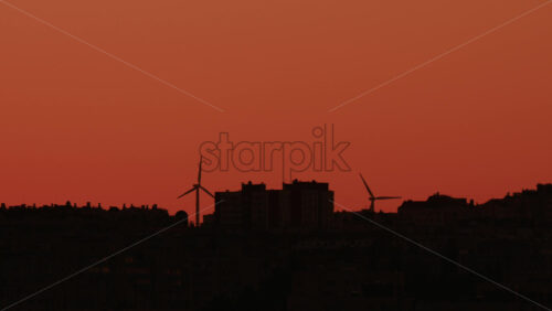 Video - Two wind turbines stand tall near city buildings under an orange sky