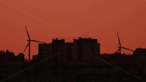 Video - Two wind turbines stand tall near city buildings under an orange sky