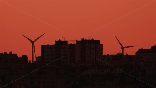 Video - Two wind turbines stand tall near city buildings under an orange sky