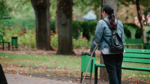 Video - A woman with crutches walks slowly along a park path surrounded by trees in daylight