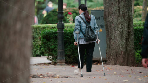 Video - A woman with crutches walks slowly along a park path surrounded by trees in daylight