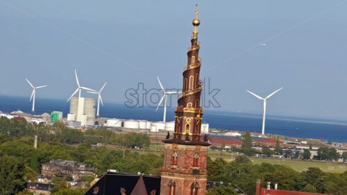 Video – Aerial drone view of the Church of Our Saviour with its golden spiral tower, set against a backdrop of wind turbines and the sea in Copenhagen, Denmark - Starpik Stock