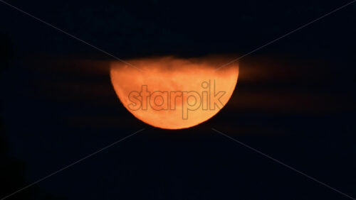 Video - A stunning orange moon appears while drifting behind clouds in a night sky.
