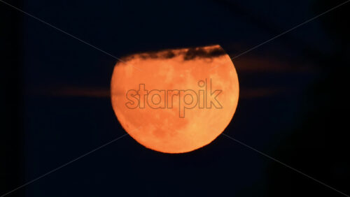 Video - A stunning full moon glows orange as it rises against a dark night sky, surrounded by wispy clouds
