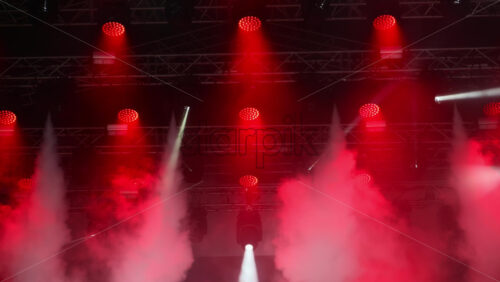 Video - Colorful lights shine down on a lively music event, illuminating the stage with red and white beams while fog adds a dramatic effect to the atmosphere.