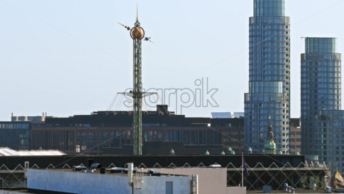 Video - Copenhagen, Denmark - August 6, 2025: Aerial drone view of Tivoli Gardens' iconic Star Flyer ride with modern skyscrapers in the background