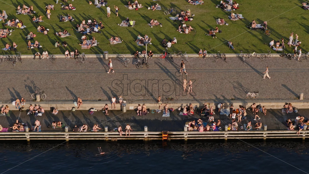 Video - Copenhagen, Denmark - August 6, 2025: Aerial view of locals gathering on the green lawn and along the harbor edge in the evening sun at Havneparken, Islands Brygge