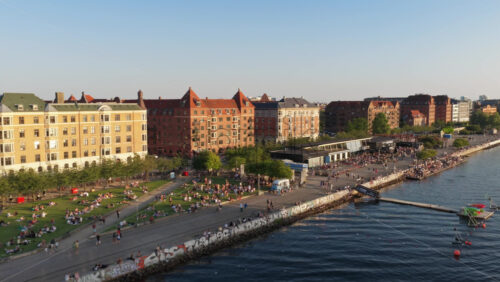 Video - Copenhagen, Denmark - August 6, 2025: Aerial drone view of the waterfront park filled with people on the grass and along the water at the Islands Brygge Park and Promenade