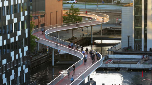 Video - Copenhagen, Denmark - August 6, 2025: Aerial drone view of red cycle bridge curving above the harbour, filled with cyclists commuting through the city