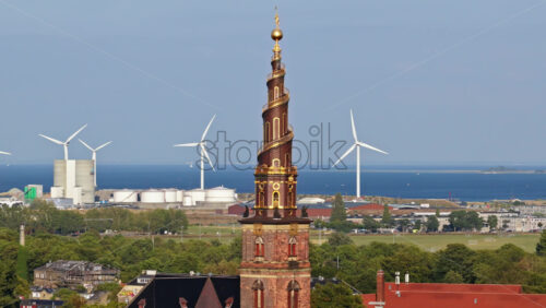 Video - Aerial drone view of the Church of Our Saviour with its golden spiral tower, set against a backdrop of wind turbines and the sea in Copenhagen, Denmark