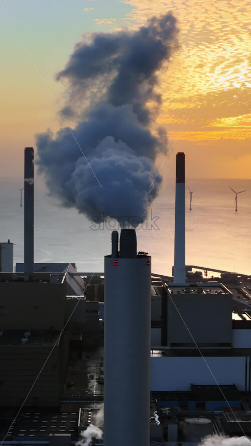 Video - Aerial drone view of Amager Bakke power plant chimney releasing smoke, with offshore wind turbines standing in the sea behind it in Copenhagen, Denmark. Vertical