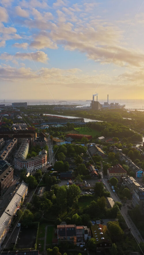 Video - Aerial drone view of Copenhagen, with a golden glow reflecting over the harbor and city rooftops, highlighting the industrial plant and wind turbines by the water. Vertical