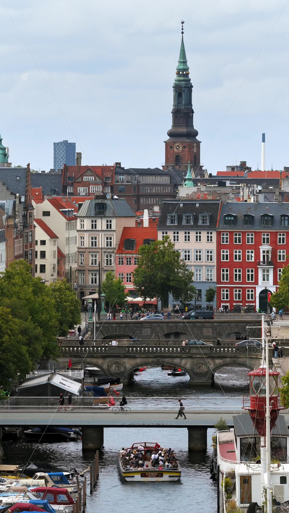 Video - Copenhagen, Denmark - August 5, 2025: Aerial drone view of a canal boat passes under bridges with the spire of Christiansborg Palace rising above central Copenhagen. Vertical