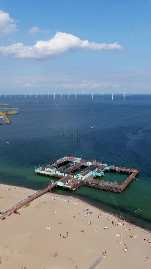 Video - Aerial drone view of Amager Beach in Copenhagen, featuring the iconic Kastrup Sea Bath with wind turbines visible in the distance across the Oresund Strait. Vertical
