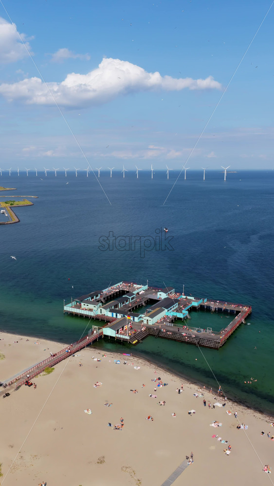 Video - Aerial drone view of Amager Beach in Copenhagen, featuring the iconic Kastrup Sea Bath with wind turbines visible in the distance across the Oresund Strait. Vertical