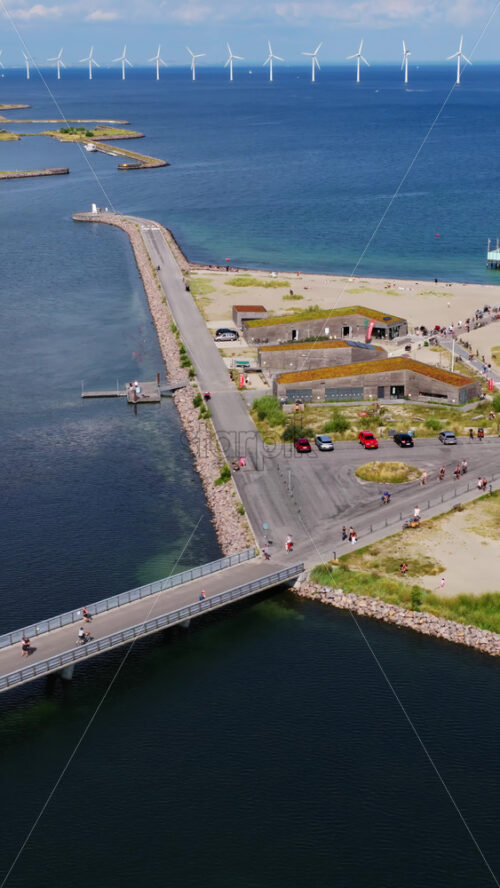 Video - Aerial drone view of Amager Strand in Copenhagen, showing the coastal road and beach with people walking along the waterfront and wind turbines in the distance. Vertical