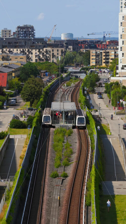 Video - Copenhagen, Denmark - August 5, 2025: Aerial drone view of two metro trains arriving at a station in Copenhagen's residential district, surrounded by modern apartments and urban greenery. Vertical