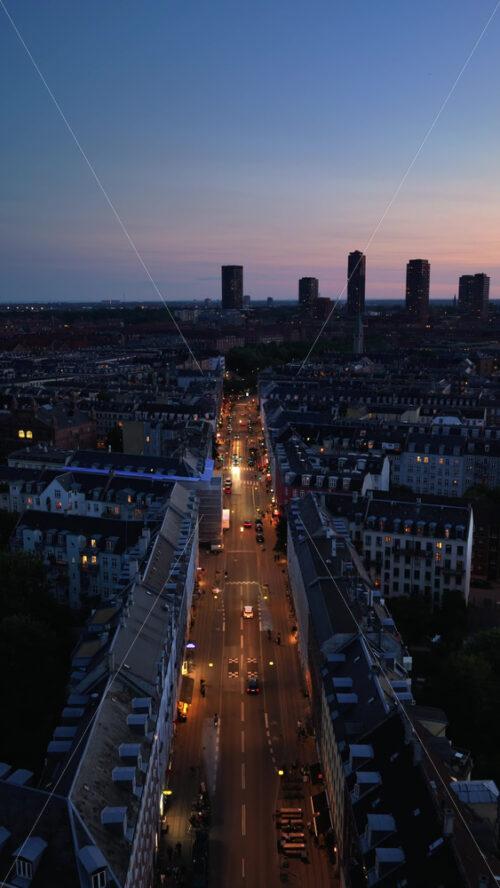 Video - Aerial drone view of Copenhagen with a long illuminated street leading into the city, while tall modern skyscrapers rise in the distance against the twilight sky. Vertical