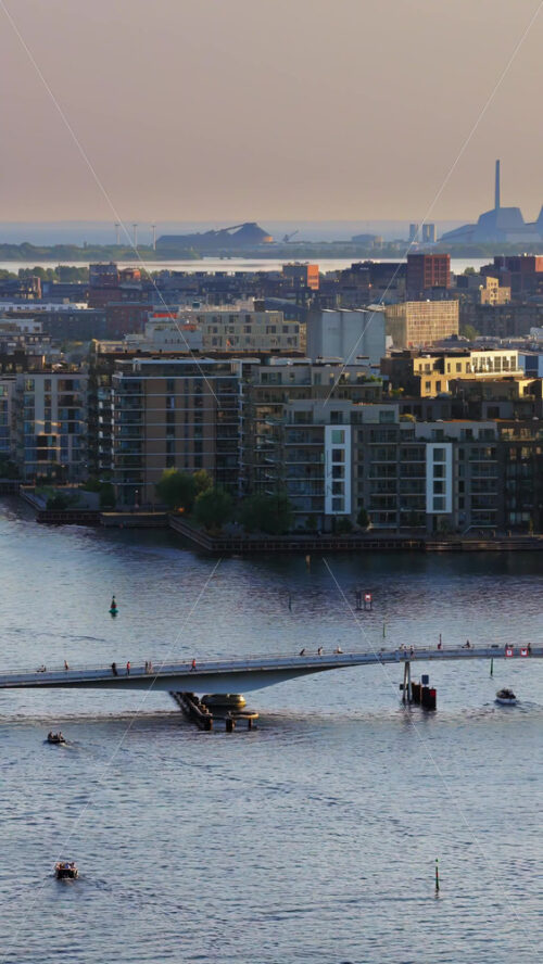 Video - Aerial drone view of the modern Lille Langebro pedestrian and cycling bridge crossing Copenhagen's inner harbor, with boats navigating the calm waters below. Vertical