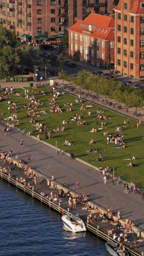 Video - Copenhagen, Denmark - August 6, 2025: Aerial view of locals gathering on the green lawn and along the harbor edge in the evening sun at Havneparken, Islands Brygge. Vertical