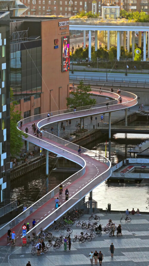 Video - Copenhagen, Denmark - August 6, 2025: Aerial drone view of red cycle bridge curving above the harbour, filled with cyclists commuting through the city. Vertical