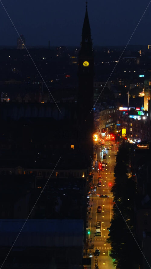 Video - Copenhagen, Denmark - August 6, 2025: Aerial drone view of Copenhagen City Hall and its clock tower, with lively city streets glowing with lights and traffic below at night. Vertical