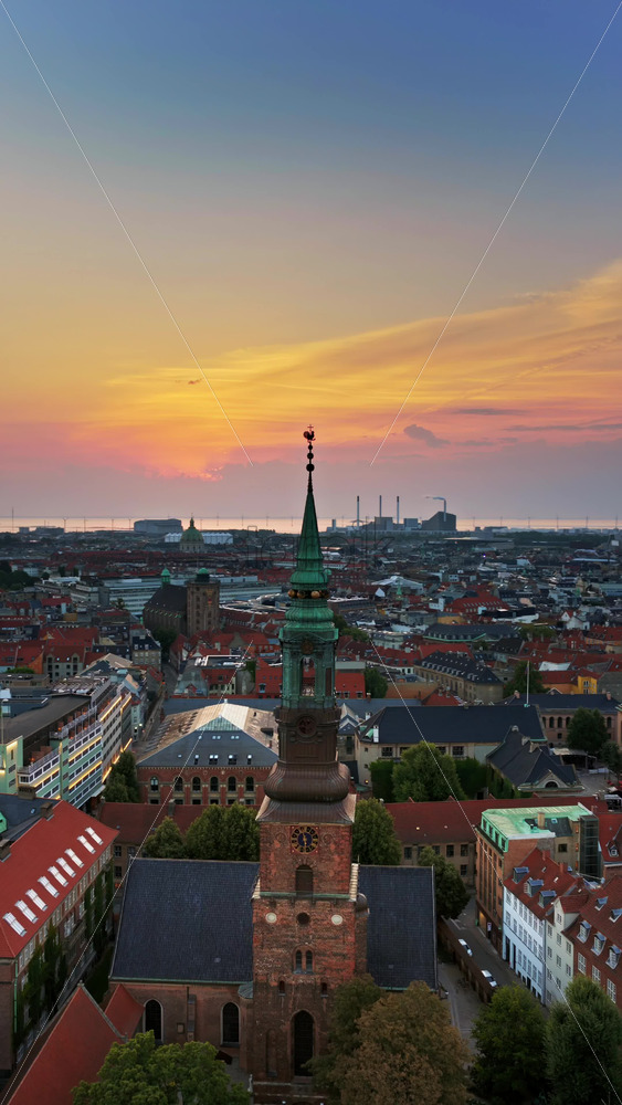 Video - Aerial drone view of St. Nicholas Church, with its green copper spire rising above the city's red rooftops at sunset in Copenhagen, Denmark. Vertical