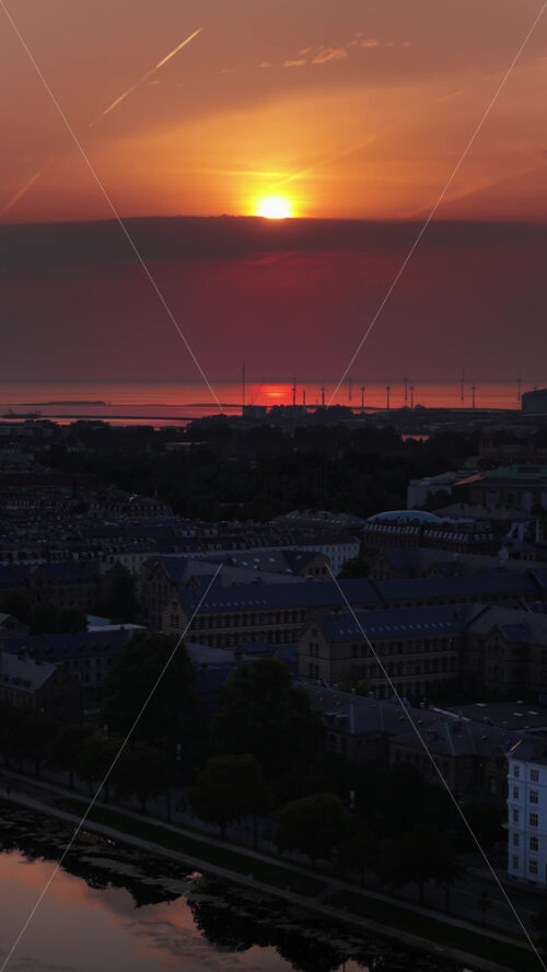 Video - Aerial drone view above Copenhagen's harbor, with the skyline and offshore wind turbines silhouetted against the glowing horizon. Vertical
