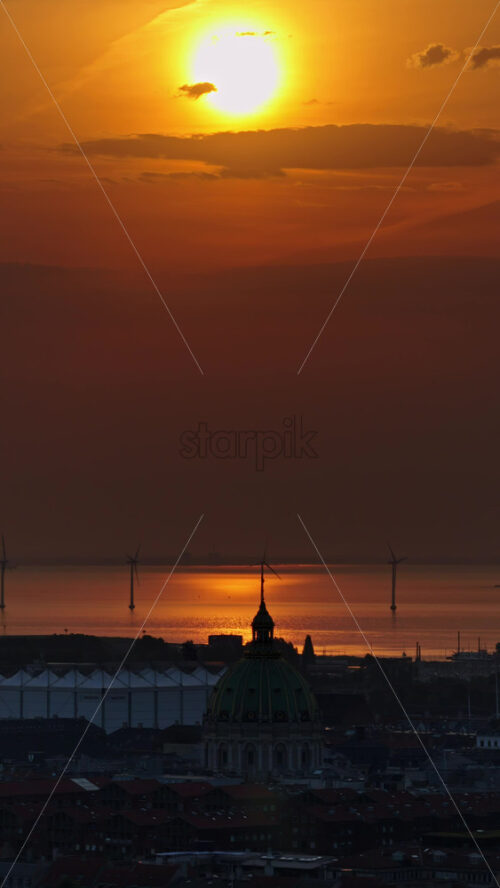Video - Aerial drone view over Copenhagen, with the Marble Church dome in the foreground and offshore wind turbines glowing in the golden light of dawn. Vertical